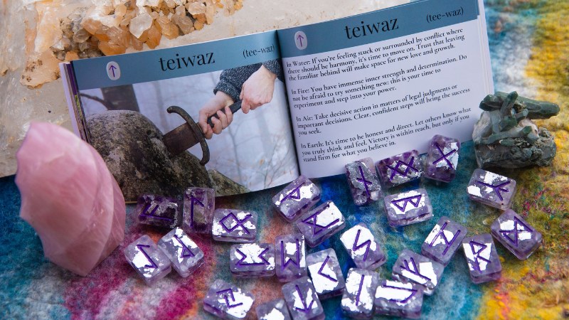 Amethyst and silver leaf runes arranged in front of an open booklet displaying the Tiwaz rune, with decorative pink Kunzite a big, local clear quartz crystal.