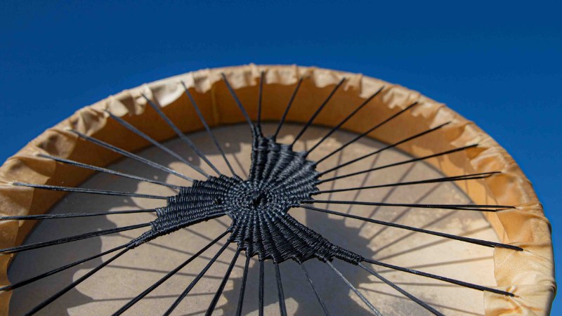 Black woven drum with three swirl pattern handle held aloft against a blue sky background.